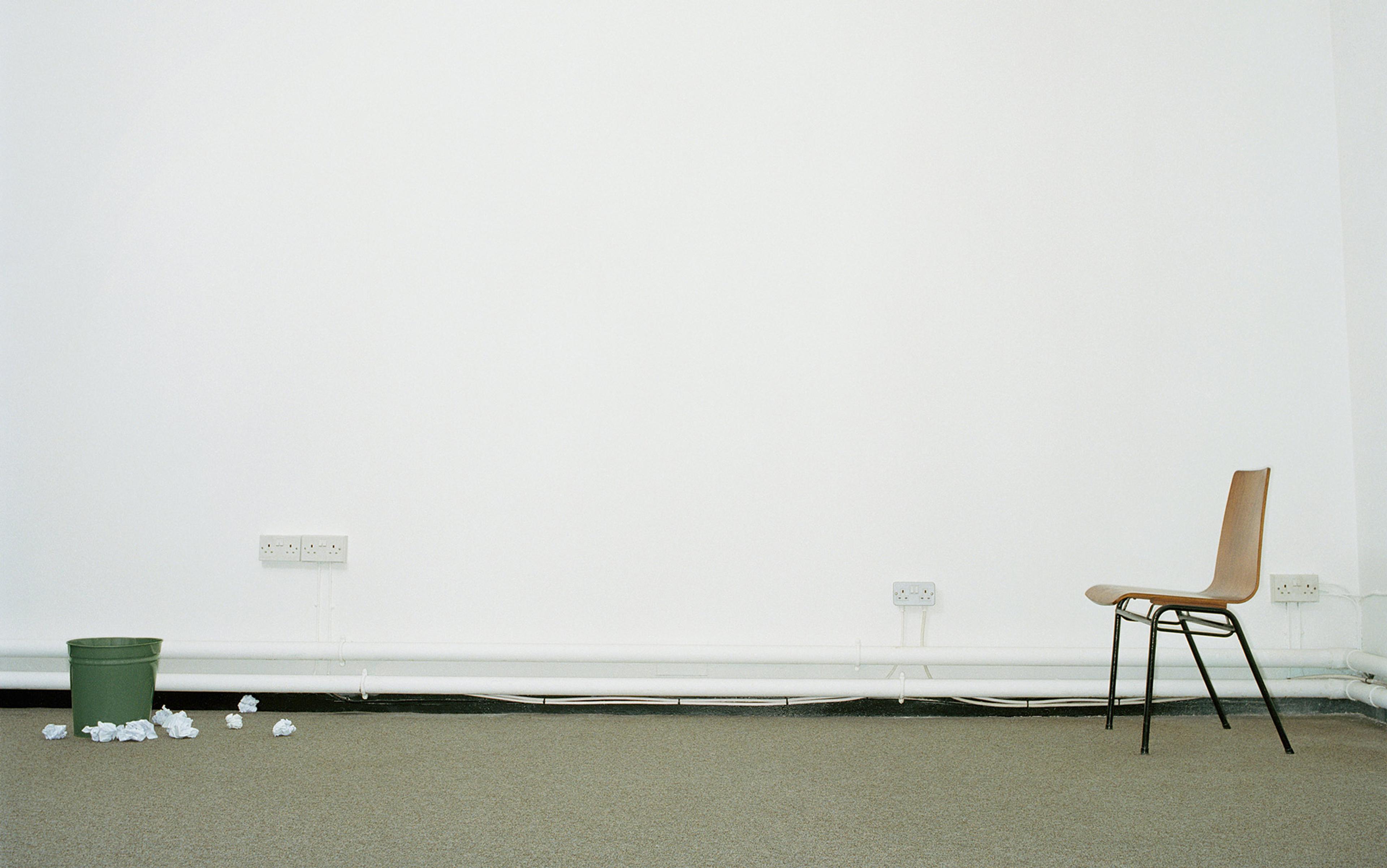 An empty room with a chair against a wall on the right, scrunched paper on the floor by a bin, and electrical sockets on the wall.