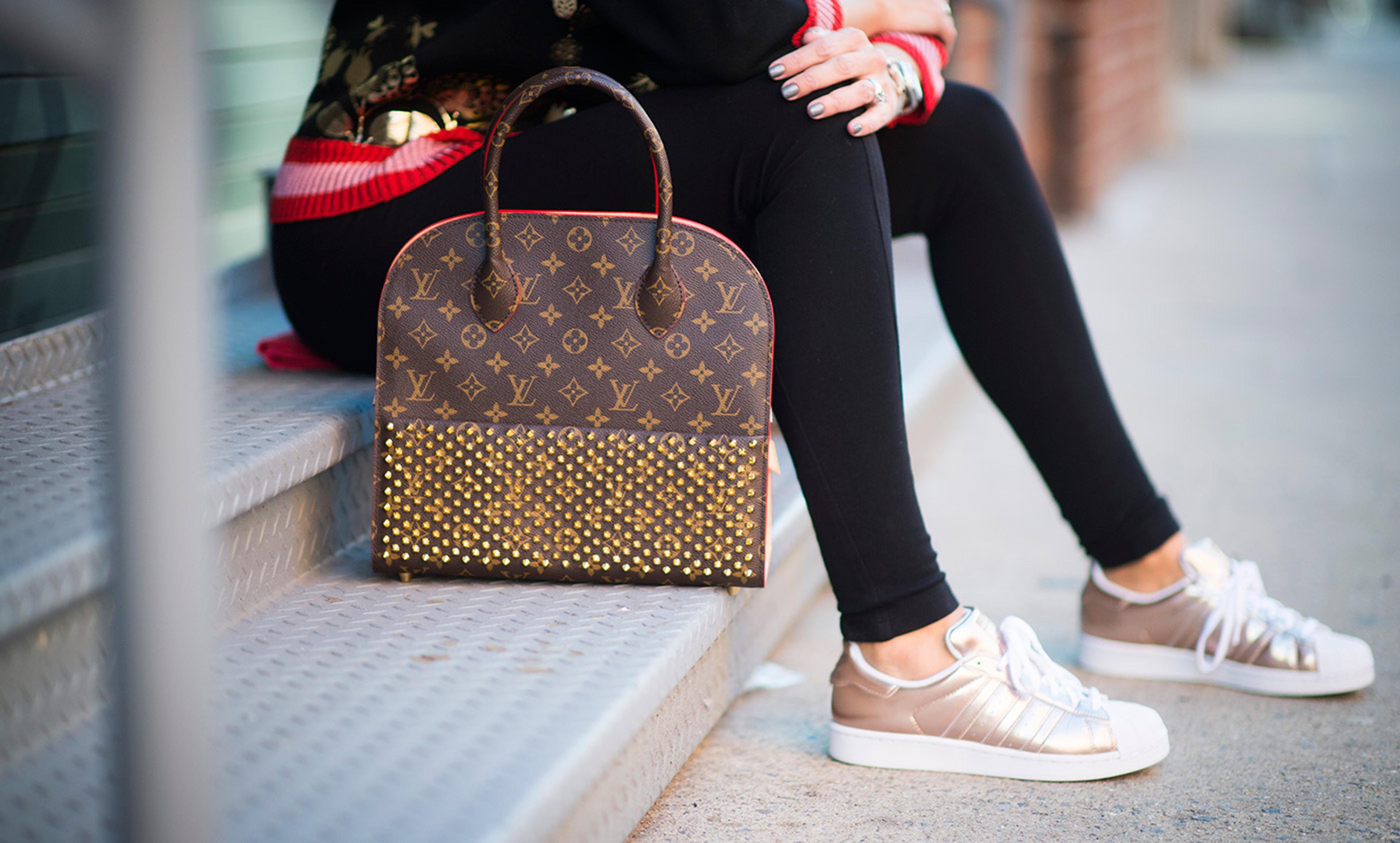 A woman sitting on stairs with a designer handbag and gold trainers, wearing black leggings and a colourful jumper.