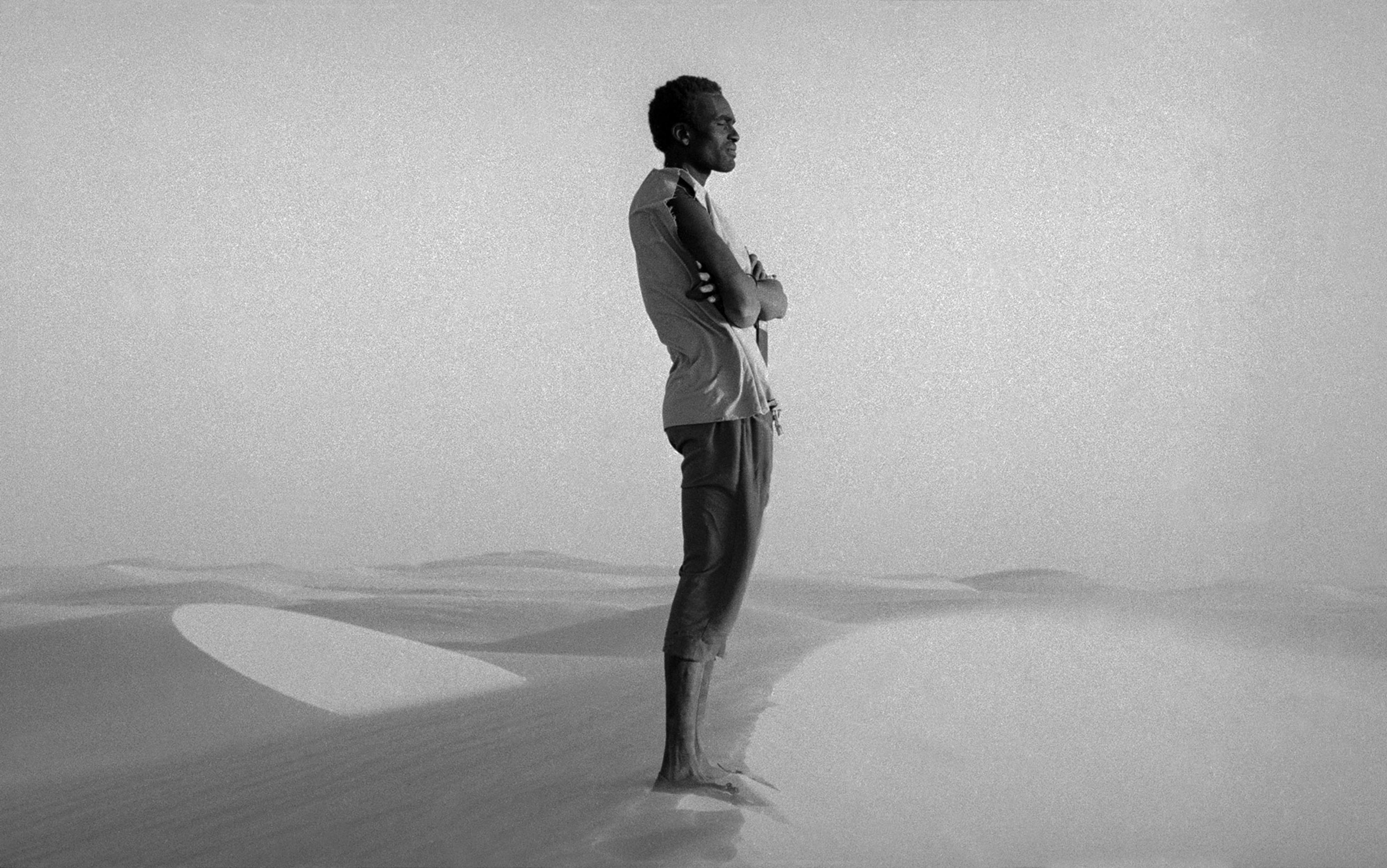 A person stands with arms crossed, looking into the distance on a dune in a vast, foggy desert. The image is in black and white.