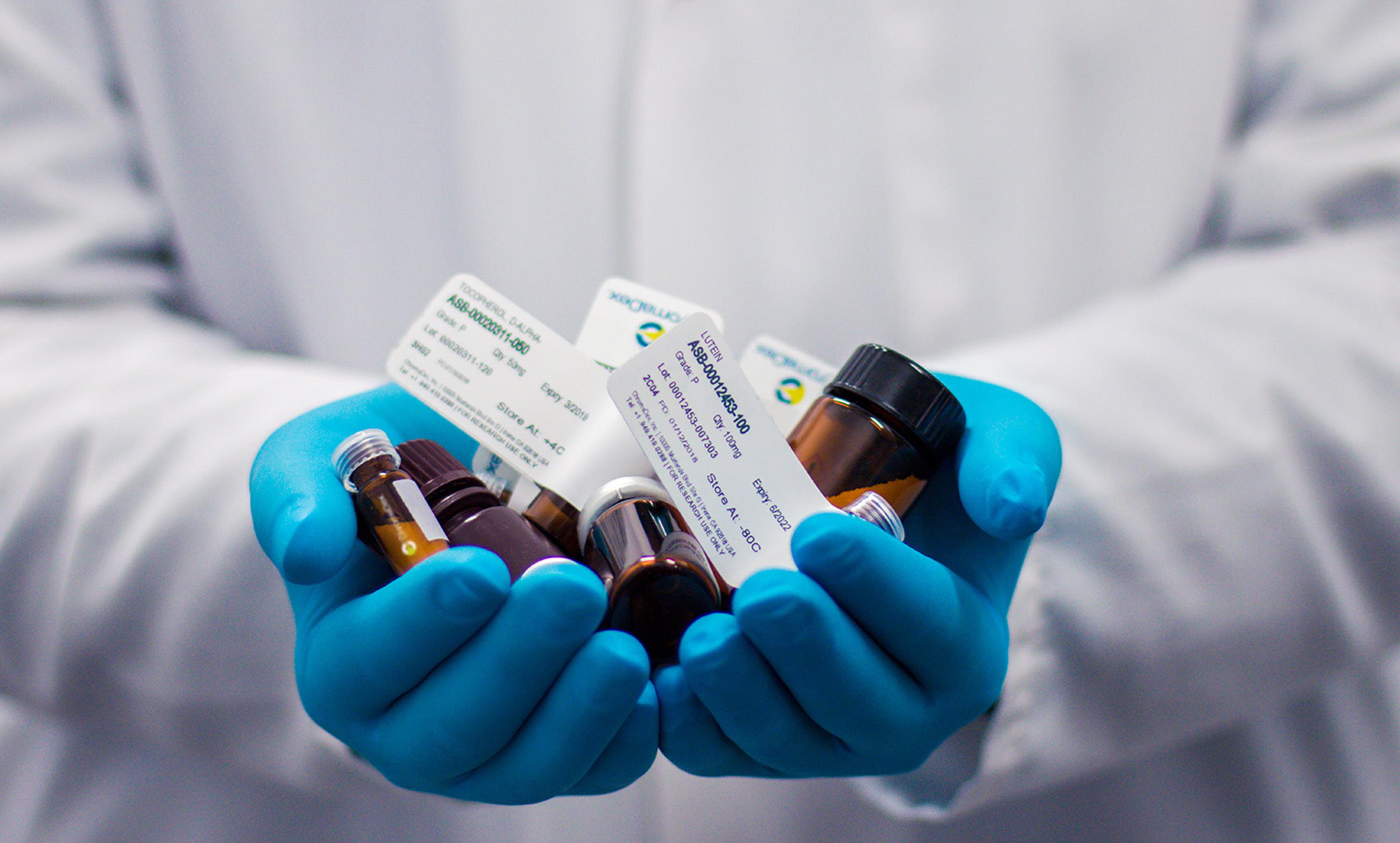 A person in a lab coat holding multiple vials and labelled containers with blue gloved hands in a laboratory setting.