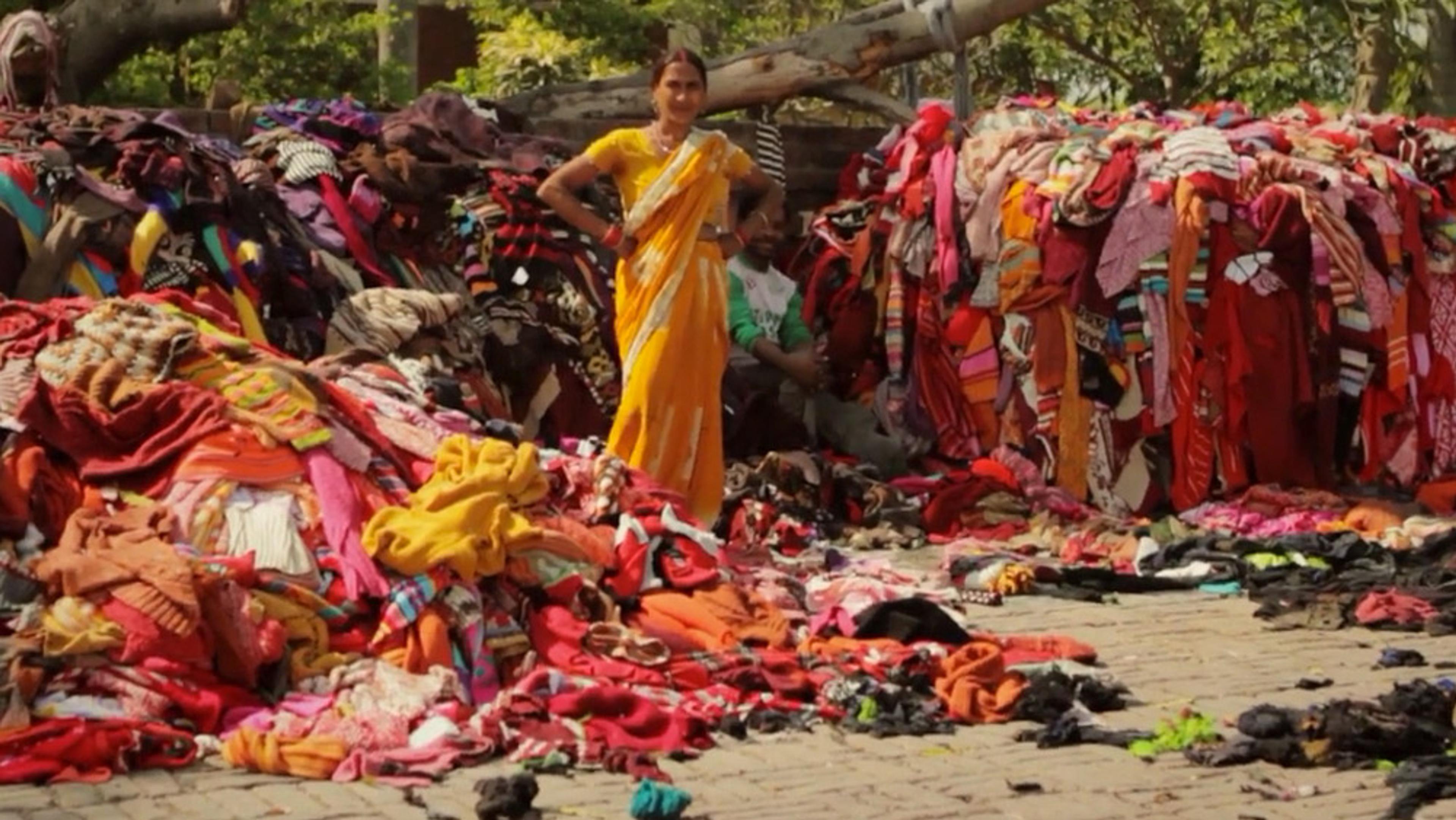 A woman in a yellow saree standing among large piles of colourful clothes outdoors, with trees in the background.