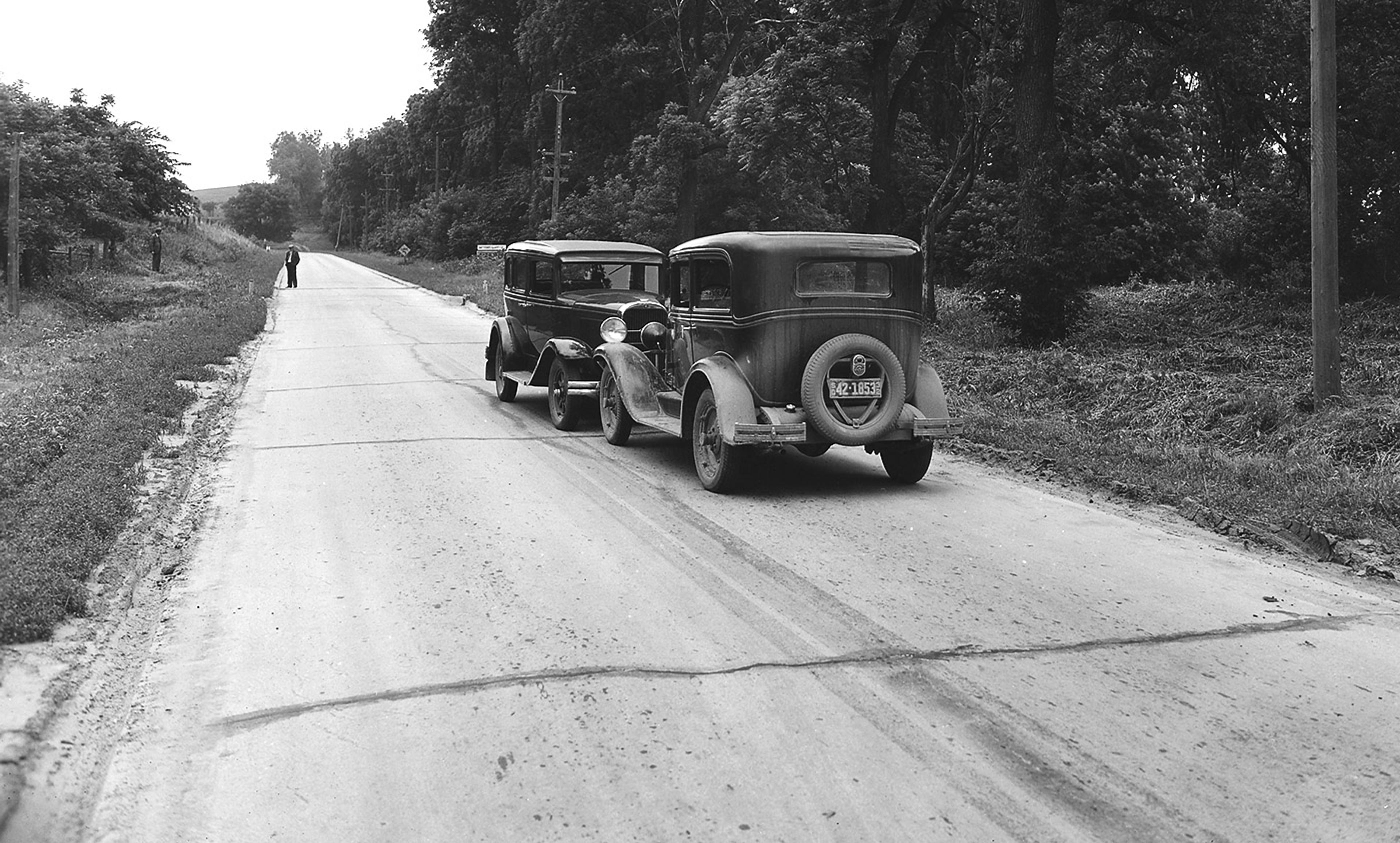 Black and white photo of two vintage cars on a rural road, lined with trees and grass, with two people walking in the distance.