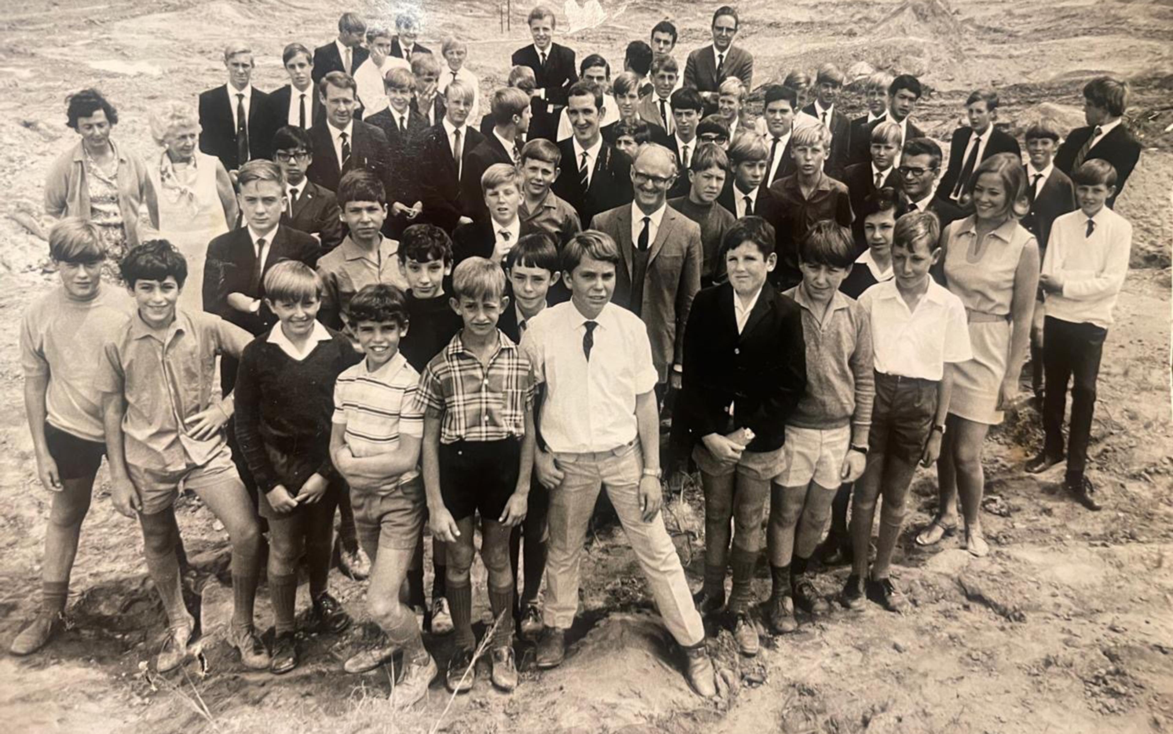 Black and white photo of a large group of boys and a few adults standing on bare ground, all dressed in 1960s attire.