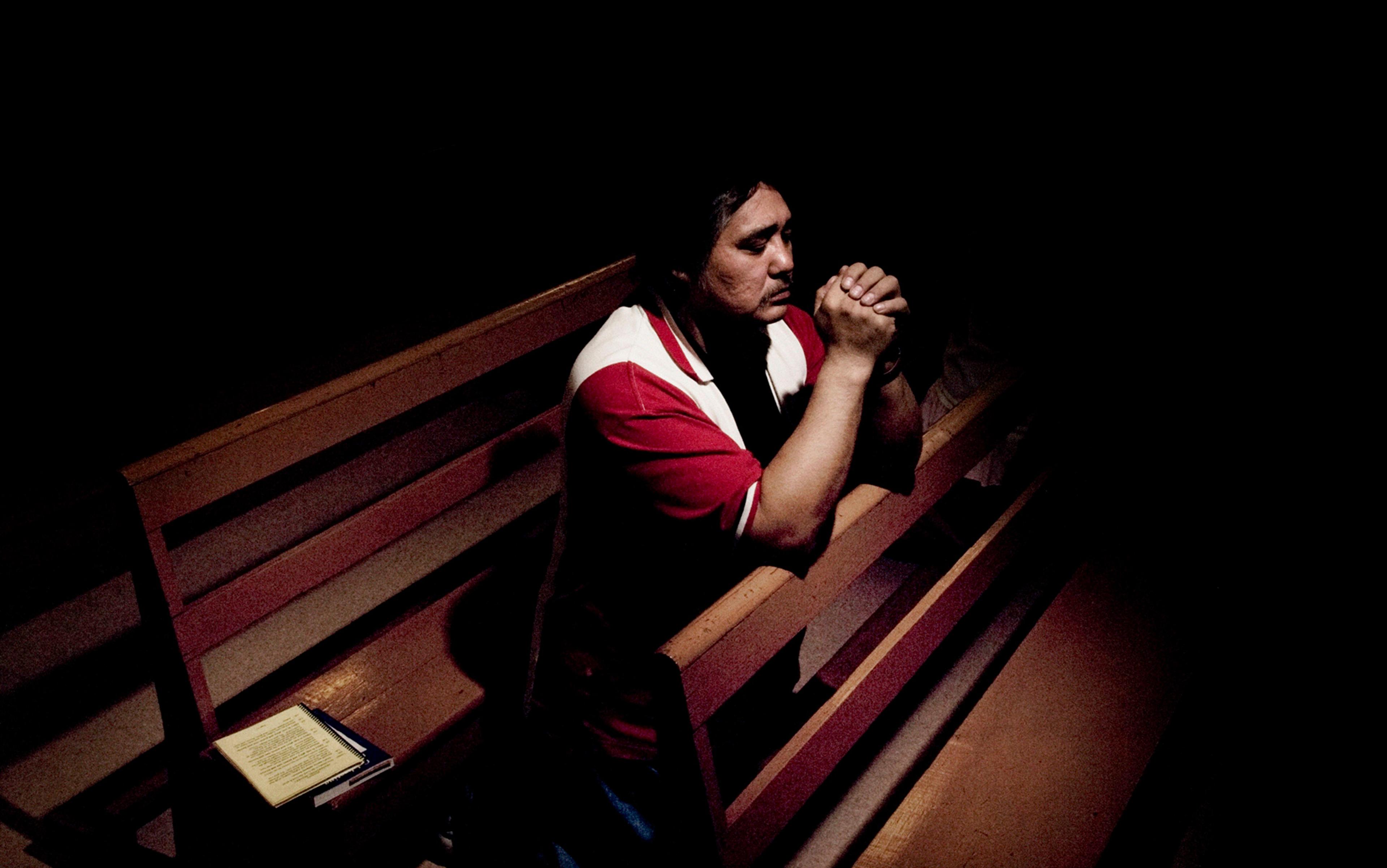 Photo of a man in a red shirt praying on a church pew, hands clasped, with a notebook beside him in dim lighting.