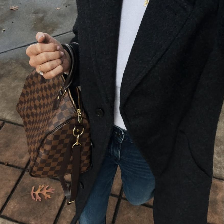 a woman in a white top and black coat holding a Louis Vuitton Damier Ebene Speedy 