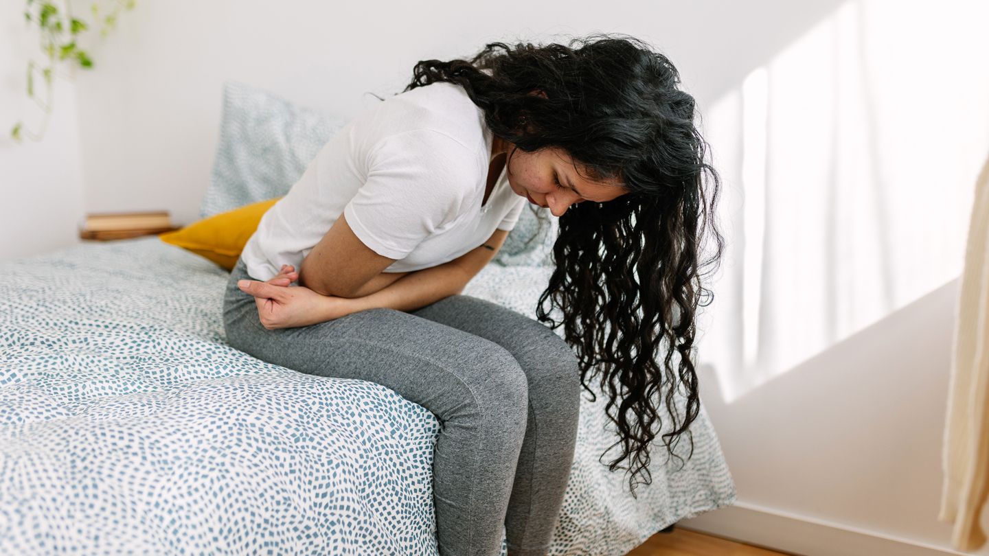 young woman clutching abdomen sitting on her bed in pain