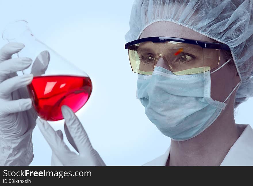 Female, caucasian lab technician with goggles and protective gear studying conical flask with red liquid. Female, caucasian lab technician with goggles and protective gear studying conical flask with red liquid