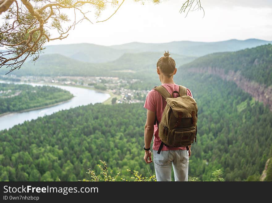 A tourist traveler with a backpack is standing on the edge of a cliff and is looking on a green valley with the river. The concept of freedom and unity with nature. A tourist traveler with a backpack is standing on the edge of a cliff and is looking on a green valley with the river. The concept of freedom and unity with nature