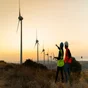 Two people in high vis jackets pointing at wind turbines in the distance at sunset