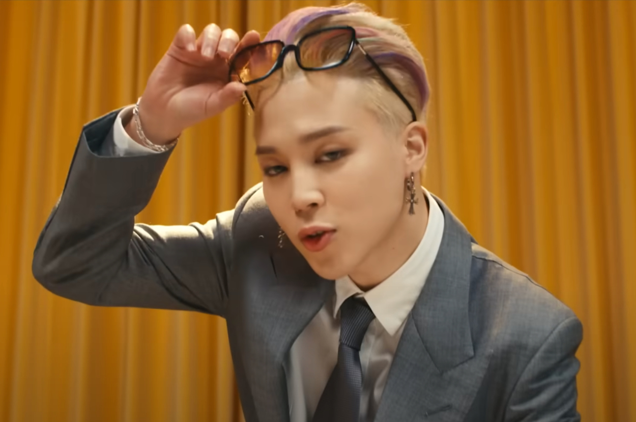 Jimin is adjusting his sunglasses, wearing a gray suit with a white shirt and dark tie, and stylish earrings, in front of a curtain backdrop