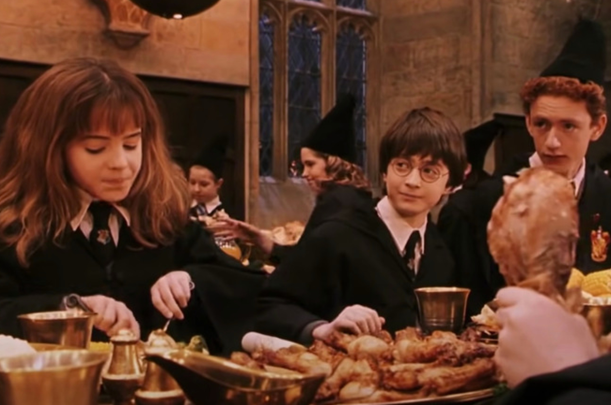 Hermione Granger, Harry Potter, and Percy Weasley, in Hogwarts uniforms, sit at a long table in the Great Hall, eating a feast. Other students are in the background