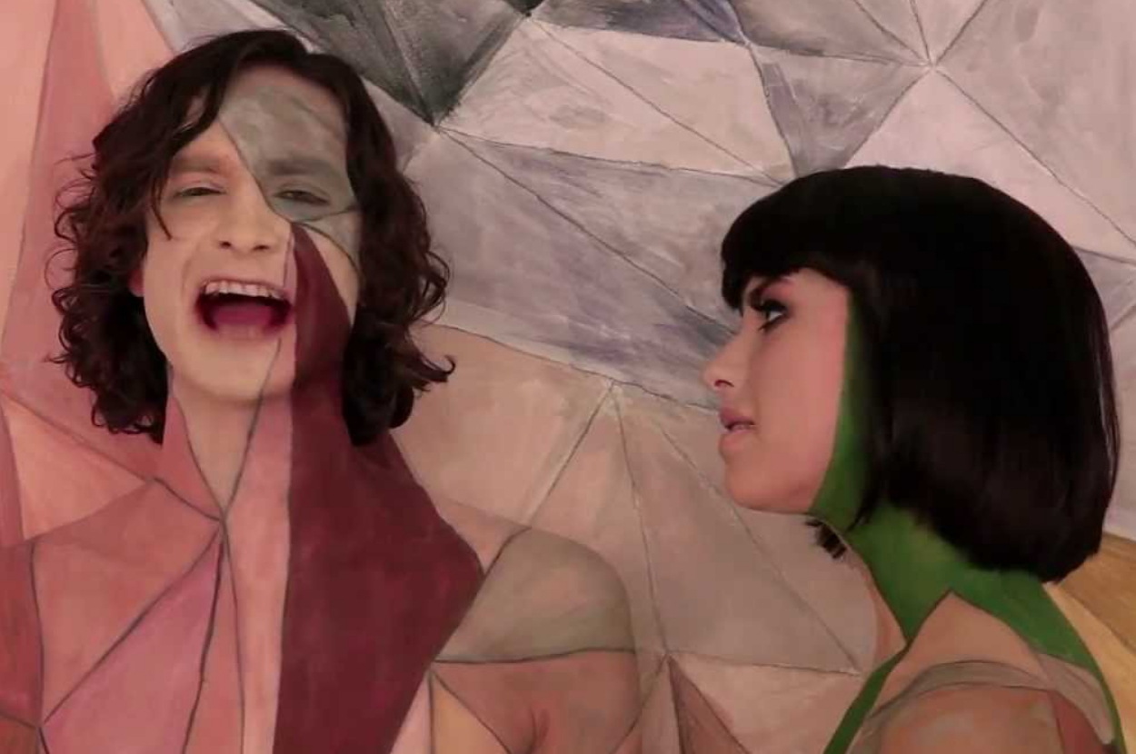 Gotye and Kimbra in a music video, with faces and bodies painted in geometric patterns resembling the background. Gotye sings while Kimbra looks on