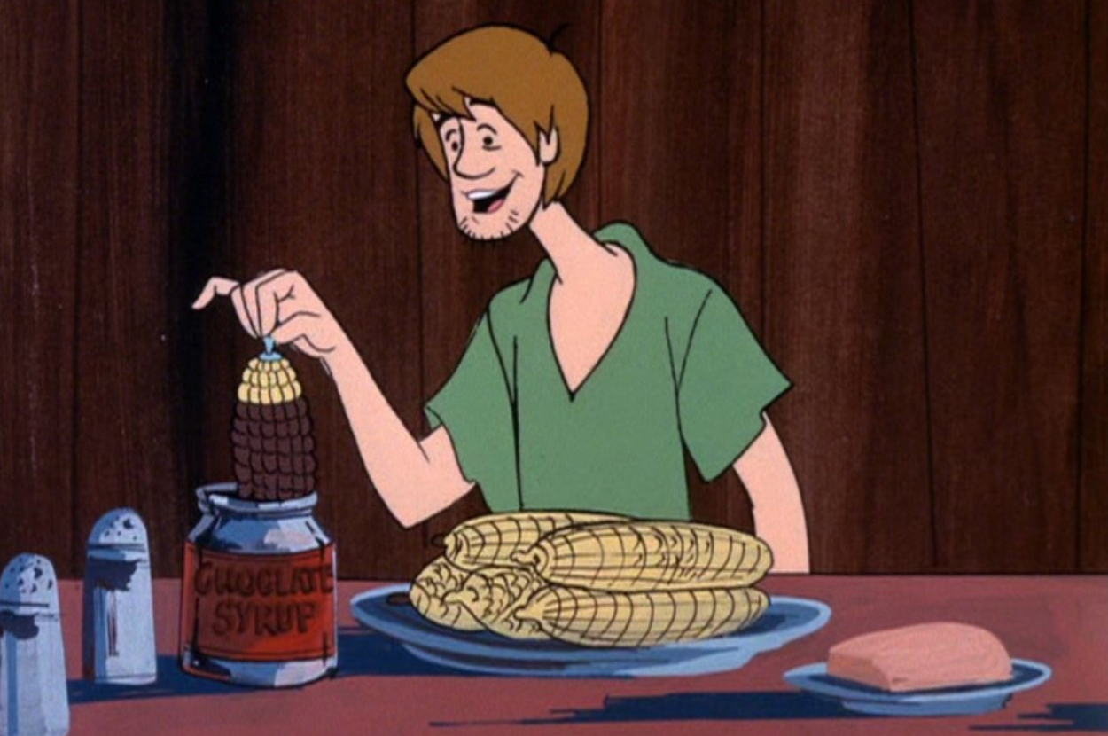 Shaggy from Scooby-Doo dips an ear of corn into a jar of chocolate syrup, with more corn and butter on the table