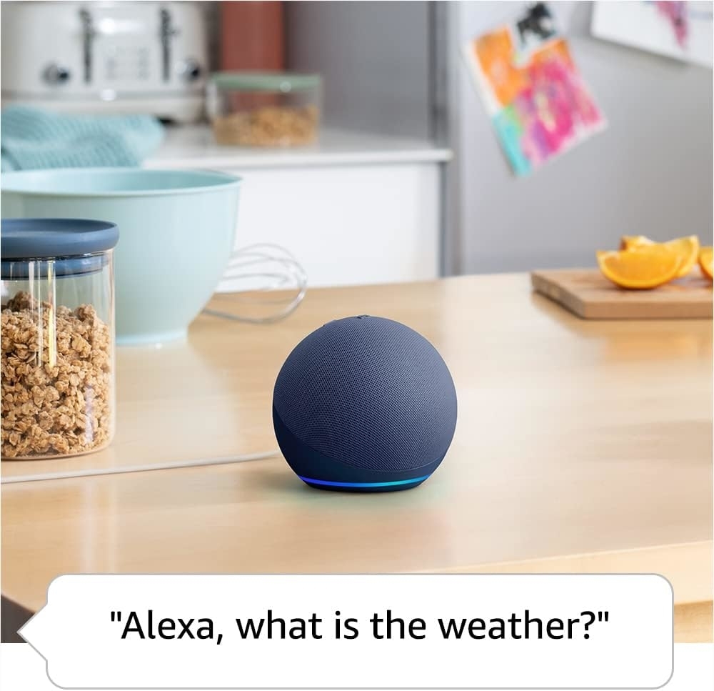 Amazon Echo Dot on a kitchen counter. The display shows the voice command, &quot;Alexa, what is the weather?&quot; nearby cereal jar and fruit in the background