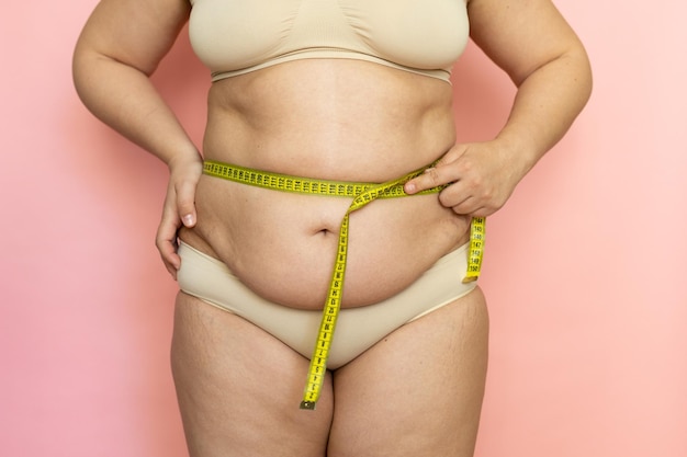 Cropped photo of naked fat and overweight woman tummy trying to\
lose weight measuring by roulette tape belly closeup