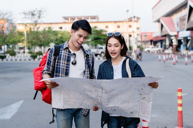 Happy asian couple tourist backpackers holding paper map and\
looking for direction while traveling, they smile with glad when\
arrived at location on paper map destination.