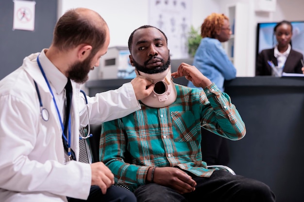 Injured african american man wearing neck collar talking to\
doctor at medical appointment in waiting room lobby. patient in\
pain with cervical foam brace after accident injury, receiving\
treatment.