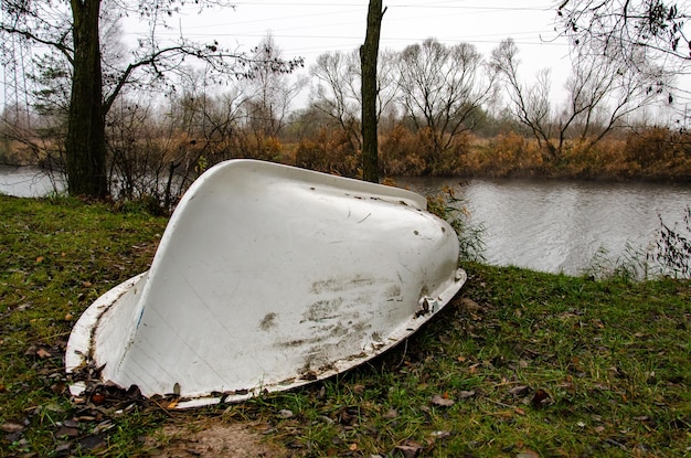 Old white boat rusting in the open dump in the forest at the\
foot of the river bank of outdated water equipment nearby grow\
large birches and other trees