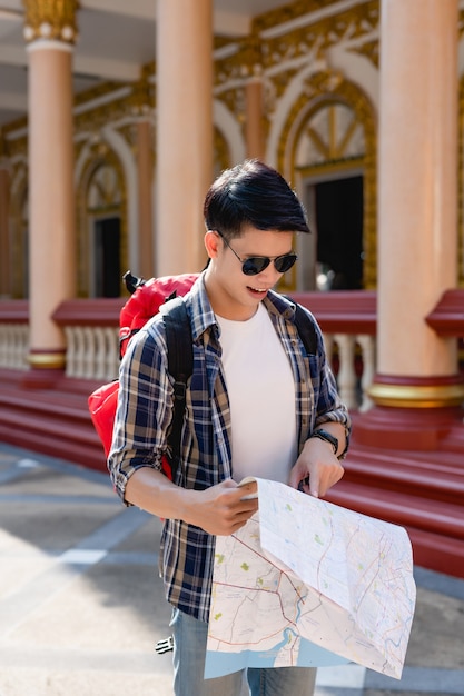 Portrait young asian backpacker male in sunglasses use finger\
point on paper map to checking direction, he smile and looking map\
at beautiful thai temple