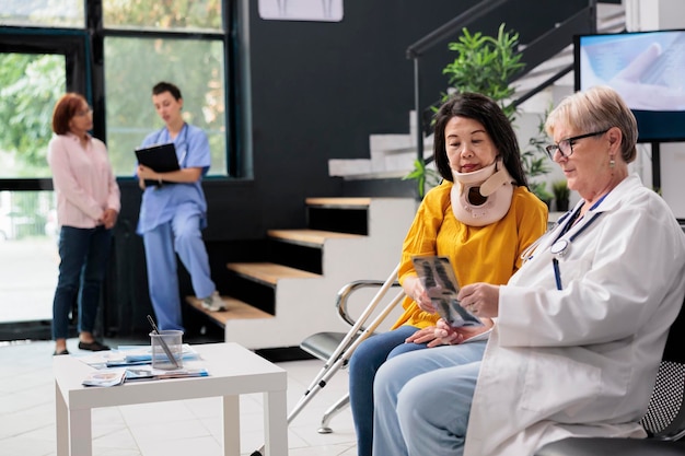 Senior doctor and patient with cervical neck collar analying\
bones radiography scan, doing checkup visit in waiting room lobby.\
asian woman with physical pain and injury having medical\
appointment.