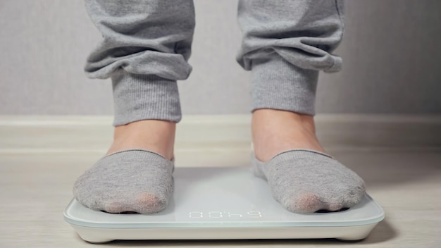 Woman in grey sweatpants and sock sstands on smart electronic\
scales and measures lost weight closeup.