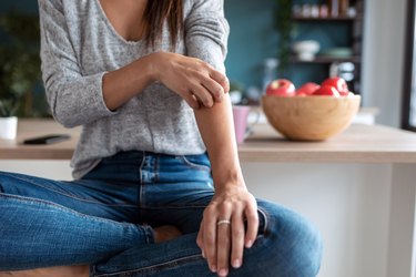 close view of a person in a grey sweater and jeans scratching their arm while sitting on a kitchen stool