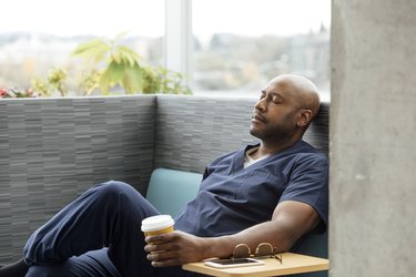 Nurse in blue scrubs sitting down, resting his eyes from getting dizzy while drinking caffeine.