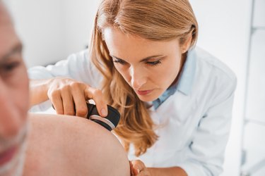 a dermatologist with long blonde hair wearing a white coat and doing a skin check on a person with white hair and a white beard