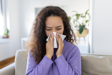 Close up of a person wearing a purple sweater with long curly brown hair sitting on a couch blowing their nose