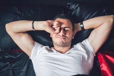 overhead photo of an adult wearing a white t-shirt waking up in bed and rubbing their eyes because of eye discharge