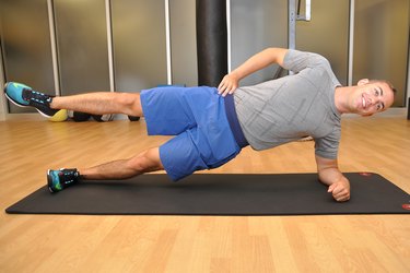 trainer and writer Henry Halse wears a gray t-shirt and blue shorts and demonstrates the Side Plank Leg Raise on a black yoga mat