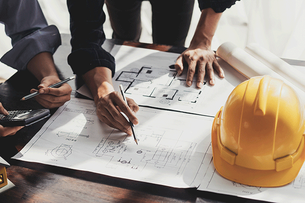 A man’s white hands are visible on a set of design plans. His left hand is planted further back on a table over top the design sheets, and his right hand is holding a pencil close to the paper. The man is wearing a black shirt with white buttons and the cuff of his left sleeve is rolled up. To the left, a woman’s hands can be seen holding a grey pencil and a silver and black calculator at the edge of the design plans. The woman is wearing a grey shirt with the cuff of one visibly rolled up.