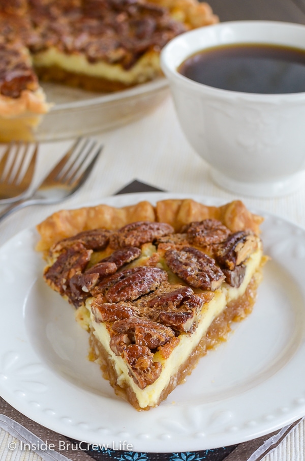 A slice of cheesecake pecan pie on a white plate with a cup of coffee behind it.