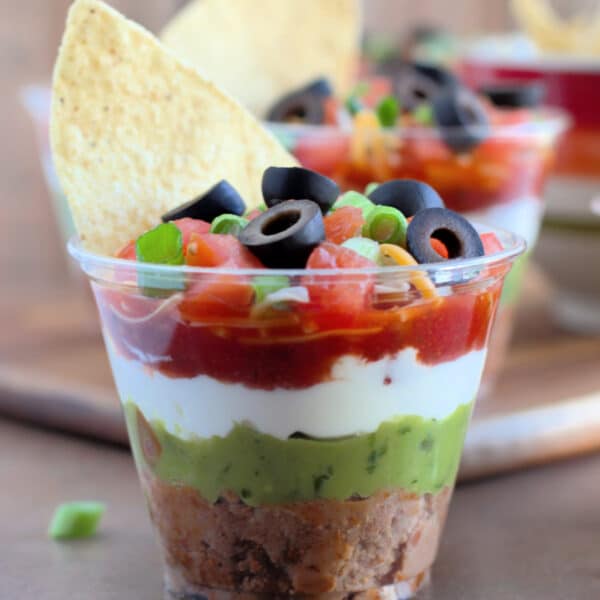 A layered seven-layer dip in a clear cup with beans, guacamole, sour cream, salsa, cheese, olives, and green onions, topped with a tortilla chip.