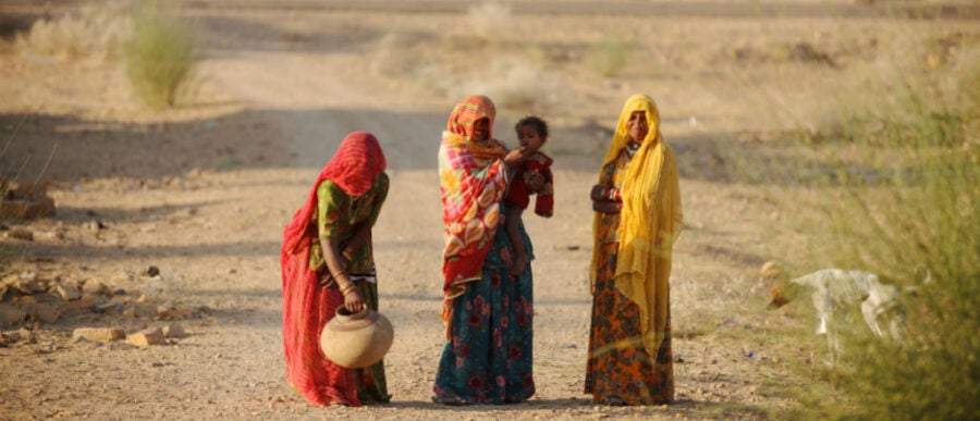 Four individuals in colorful traditional clothing stand on a dirt path in a rural landscape. One person holds a small child, and another carries a clay pot. Bushes and a dog are nearby.