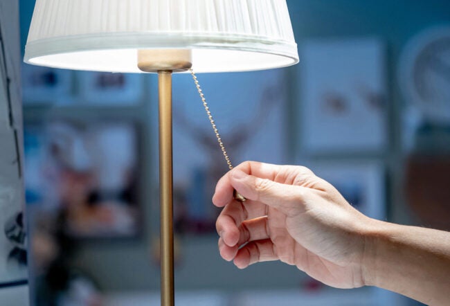 A hand pulling the chain of a lit table lamp with a pleated white shade. The background is softly blurred, suggesting an indoor setting.