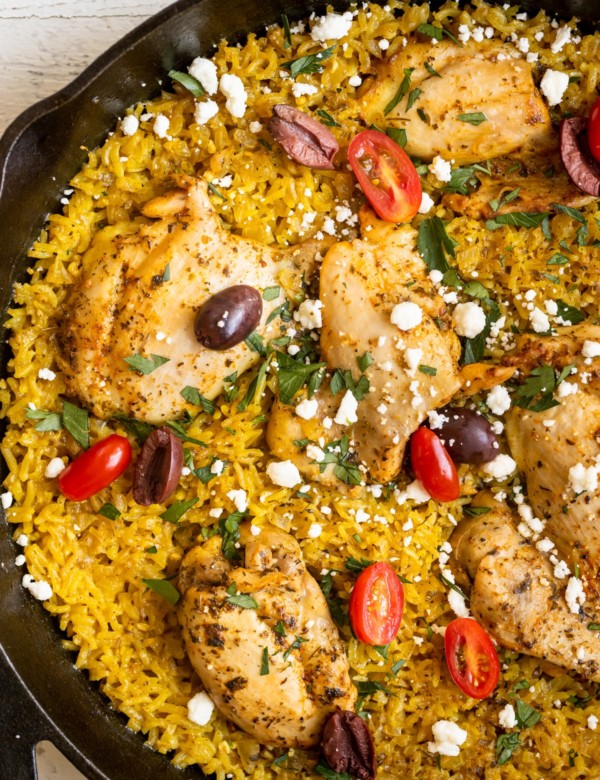 chicken thighs and yellow rice in a cast iron skillet garnished with feta, parsley, tomatoes and kalamata olives