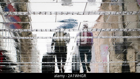 Blurry reflection silhouette on wet city street of two tourist  people walking under umbrella and  pulling traveling suitcases in the rainy autumn day Stock Photo