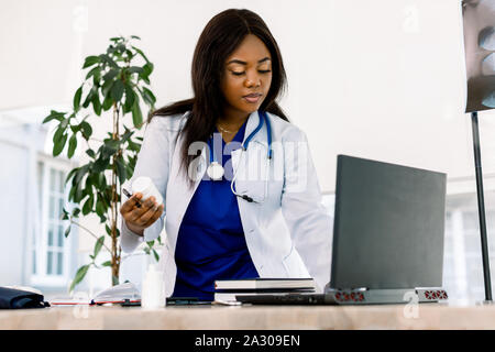 Black female doctor at work in an office, looking to laptop. Beautiful African American woman doctor or nurse working on a laptop computer in modern Stock Photo