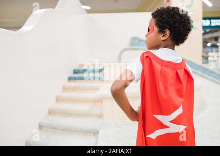 Back view of little African boy in costume of superman standing on play area Stock Photo