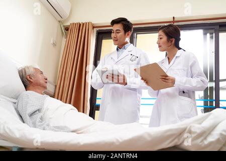 two young friendly asian doctors talking to senior asian male patient at bedside in hospital Stock Photo