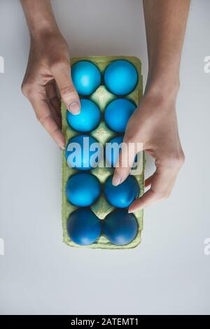 Above view background female hand arranging composition of painted blue Easter eggs in crate on white, copy space Stock Photo