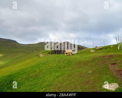 Faroe Islands. Slopes of hills rising above the Gjógv village. Wooden bench in the background. Cloudy sky. Stock Photo