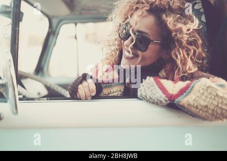 Cheerful happy beautiful caucasian young woman looking and smiling out of the window from old vintage van bus with sunglasses - travel driving concept for cute attractive girl with curly hair Stock Photo