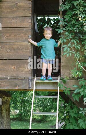 Small boy stands on stairs on treehouse in green garden. Summer time Stock Photo