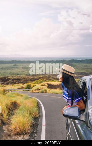 A young female wearing a hat leaning out of a car window with a beautiful landscape in the background Stock Photo