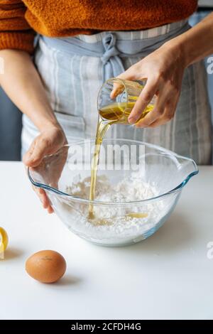 Crop female pouring olive oil from glass into large bowl with flour while preparing dough at white table in kitchen Stock Photo