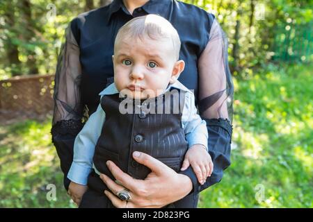 Portrait of grumpy and cute little infant wearing formal clothing sitting in mothers arms and posing for camera isolated against plain background Stock Photo