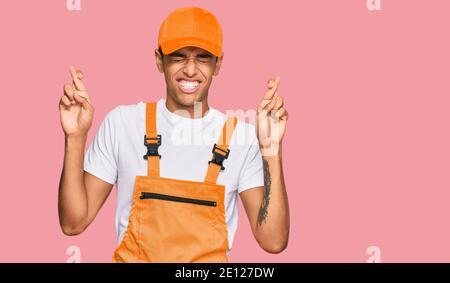 Young handsome african american man wearing handyman uniform gesturing finger crossed smiling with hope and eyes closed. luck and superstitious concep Stock Photo