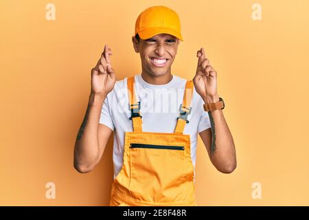 Young handsome african american man wearing handyman uniform over yellow background gesturing finger crossed smiling with hope and eyes closed. luck a Stock Photo